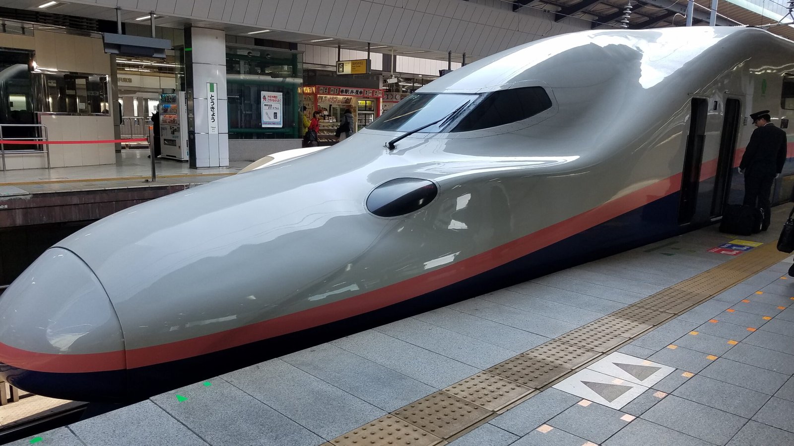Shinkansen bullet train at Tokyo Station platform in Japan; sleek silver train with a red lower stripe and blue undercarriage, with passengers and vending machines visible in the background.