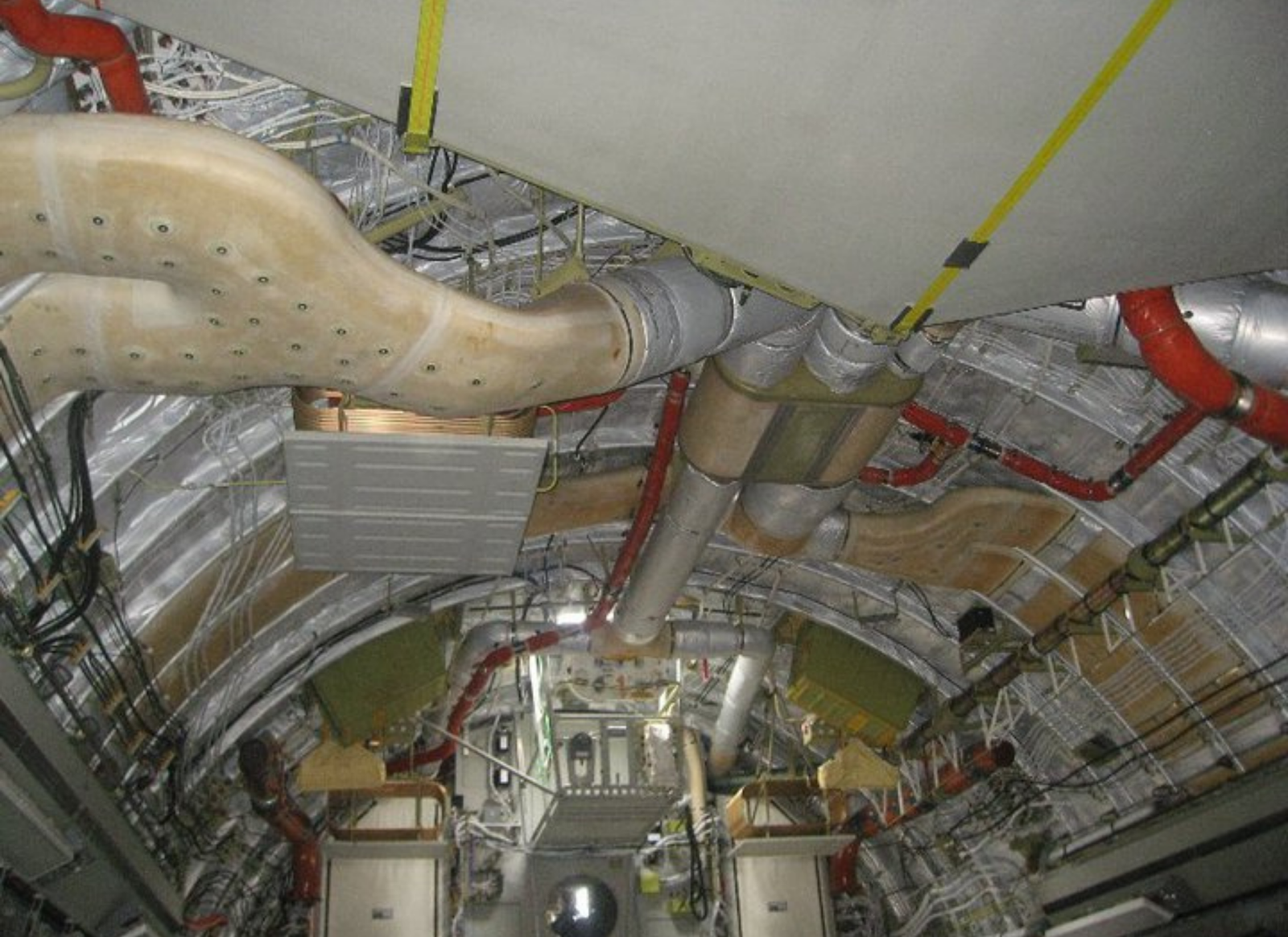 Interior of an aircraft fuselage area showing an intricate network of insulated ducts and pipes, electrical wiring, and conduit along curved metal walls, with yellow straps and green equipment boxes.