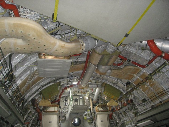 Interior of an aircraft fuselage area showing an intricate network of insulated ducts and pipes, electrical wiring, and conduit along curved metal walls, with yellow straps and green equipment boxes.
