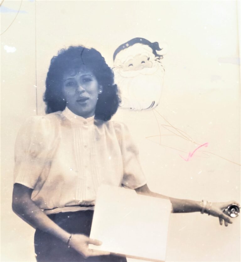 Vintage photo of a woman in a white blouse, standing indoors, holding a folder with her right hand, her left hand on a doorknob. The door is decorated with a Santa Claus drawing, 1980s holiday setting.