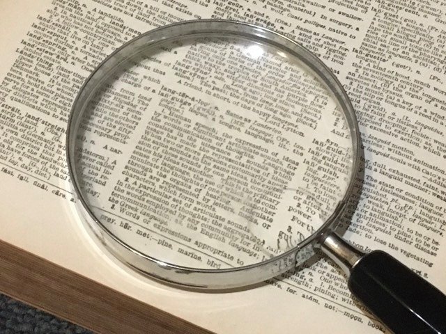 Close-up of a black-handled magnifying glass resting on an open dictionary page, magnifying the small print and word definitions.