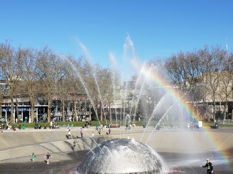 Seattle Center fountain with a large dome-shaped central jet shooting water into the air, forming a rainbow across the spray as families and individuals walk, play, and relax along a tree-lined urban plaza with benches and buildings in the background.