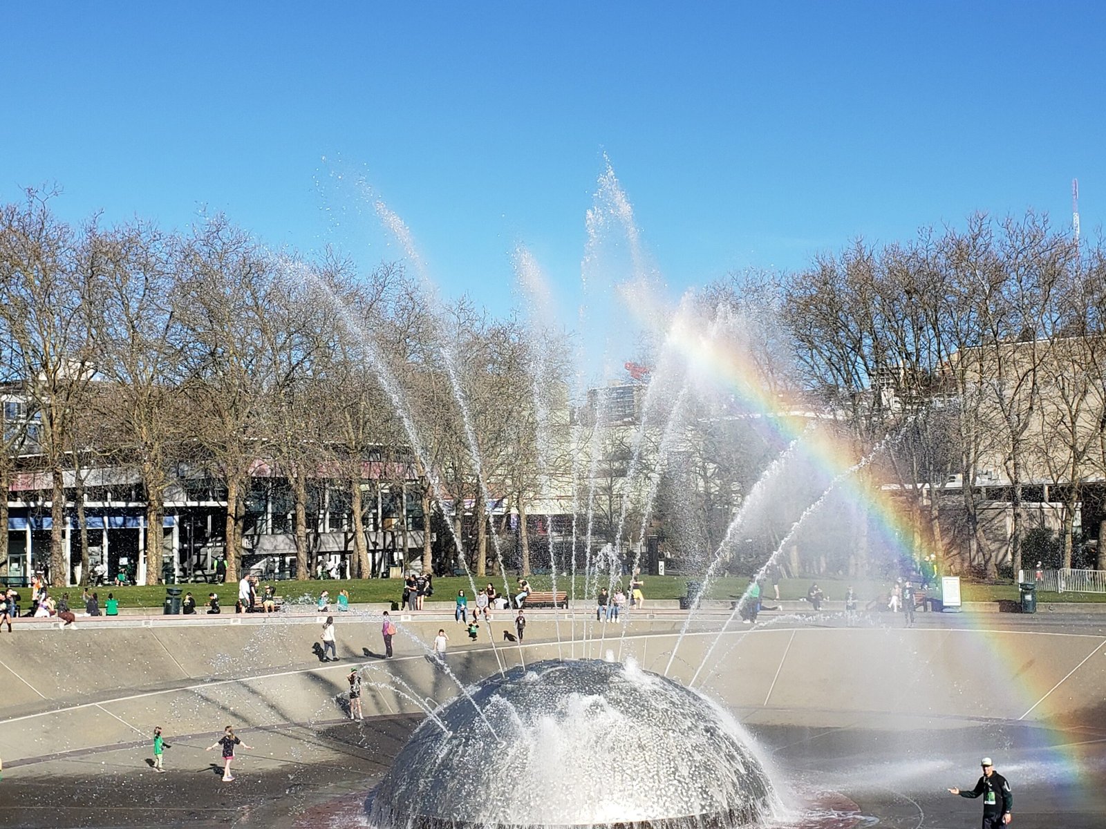 Seattle Center fountain with a large dome-shaped central jet shooting water into the air, forming a rainbow across the spray as families and individuals walk, play, and relax along a tree-lined urban plaza with benches and buildings in the background.