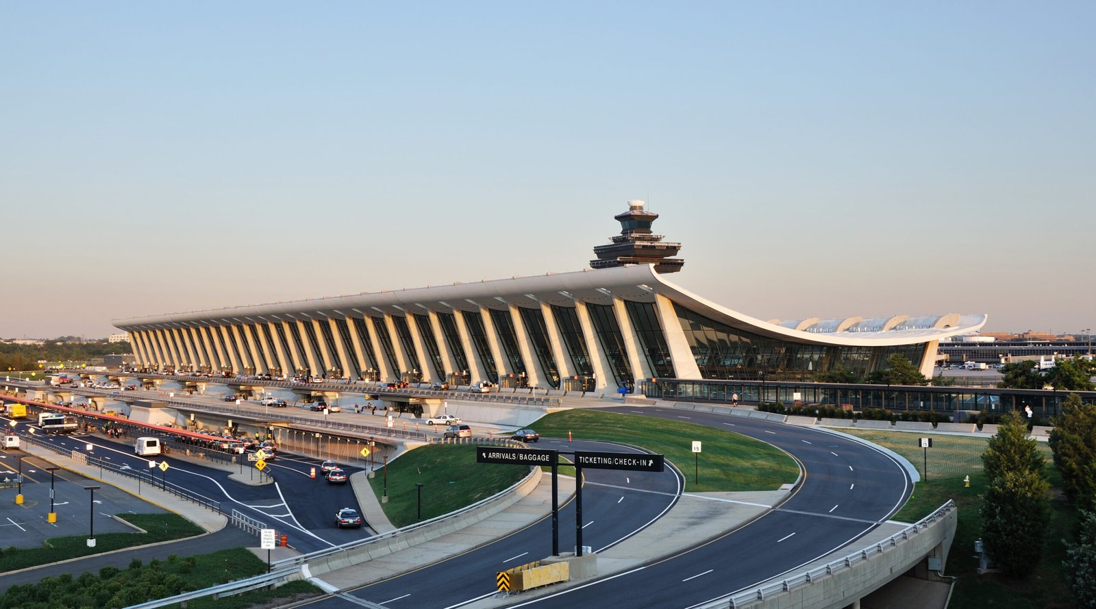 Exterior view of a modern international airport terminal (Washington Dulles Airport, D.C.) with a distinctive stacked control tower, a sweeping curved white roof, glass walls, and busy roadways with cars and signs for arrivals/baggage and ticketing/check-in.