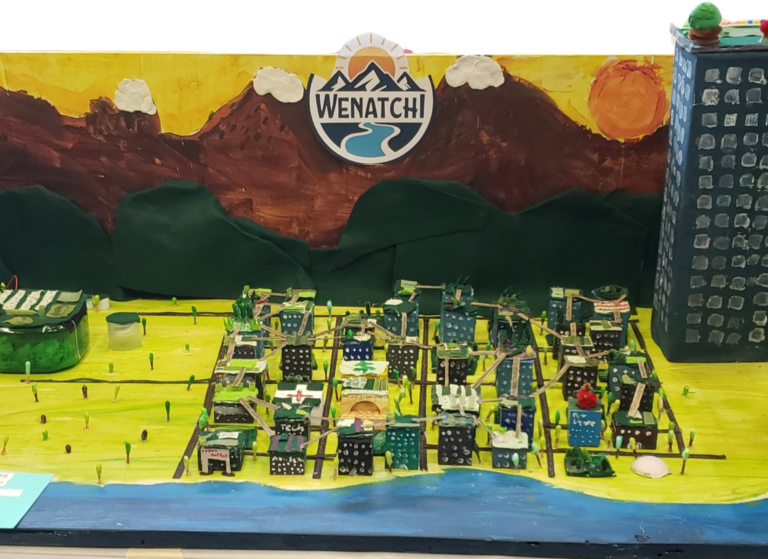 City of Wenatchi diorama on a bright yellow display featuring a grid of tiny painted buildings connected by sticks, green hills and brown mountains capped with snow rising in the background. A blue river, representing the Columbia River, along the front, and a tall blue building on the right, with a round Wenatchi logo showing mountains and a sun in the center.